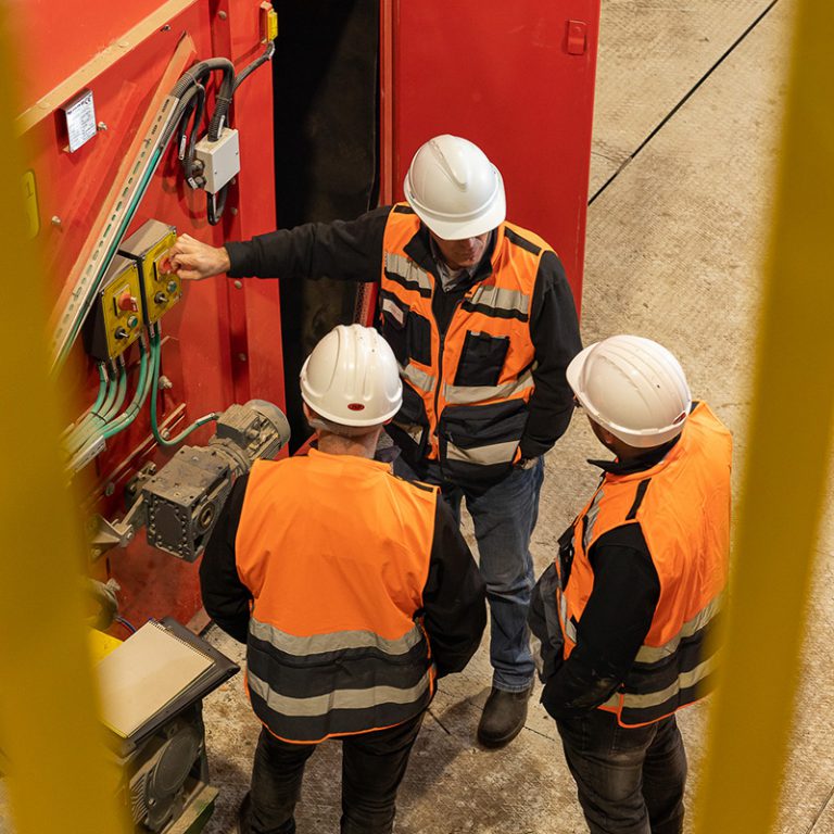 שלושה גברים חובשי קסדות ואפודים זוהרים עומדים ליד לוח בקרה של מכונה